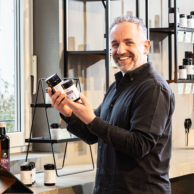 Enrico Mandl hält zwei NUI Noni Produktdosen in der Hand und steht in einem Raum mit Regalen.